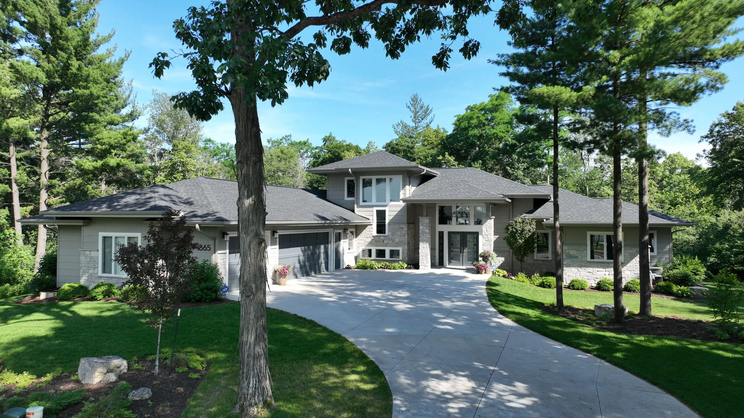 Modern single-story house with gray exterior, stone accents, large driveway, and surrounded by trees and green lawn.